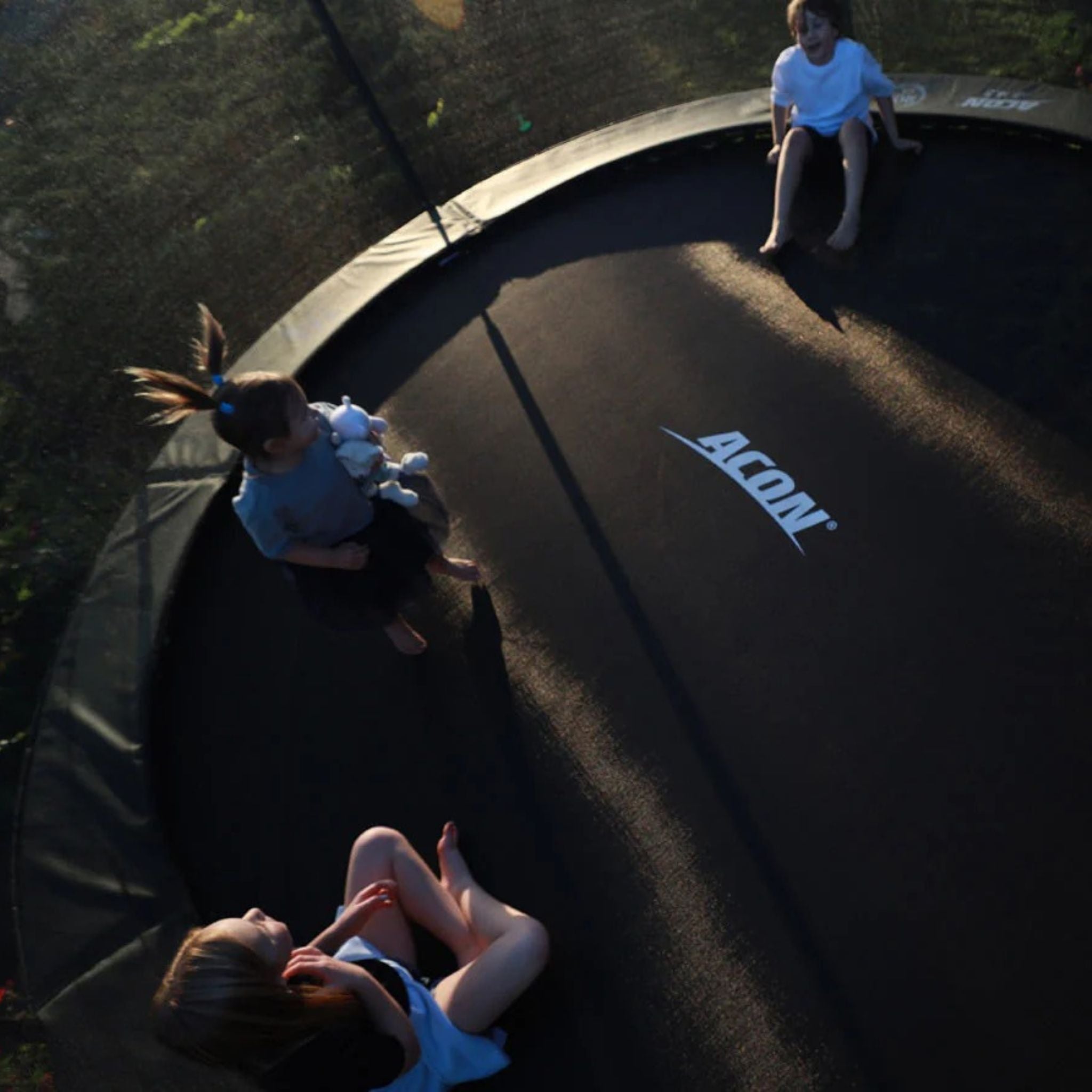 Drei Kinder sitzen auf einem Trampolin in der Sonne und genießen das Spielen im Freien.