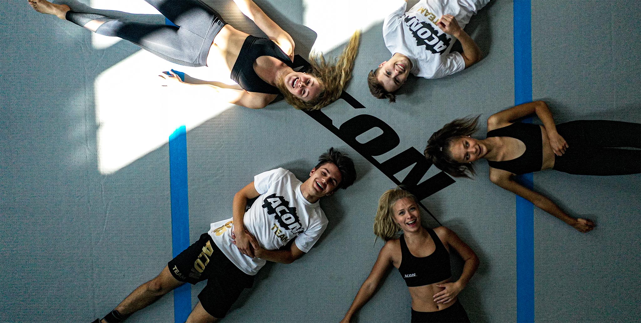 5 Acon-Teammitglieder liegen auf einer großen Airtrack. Auf dem Bild aus der Vogelperspektive sind drei junge Frauen und zwei Männer zu sehen. Alle fünf lächeln