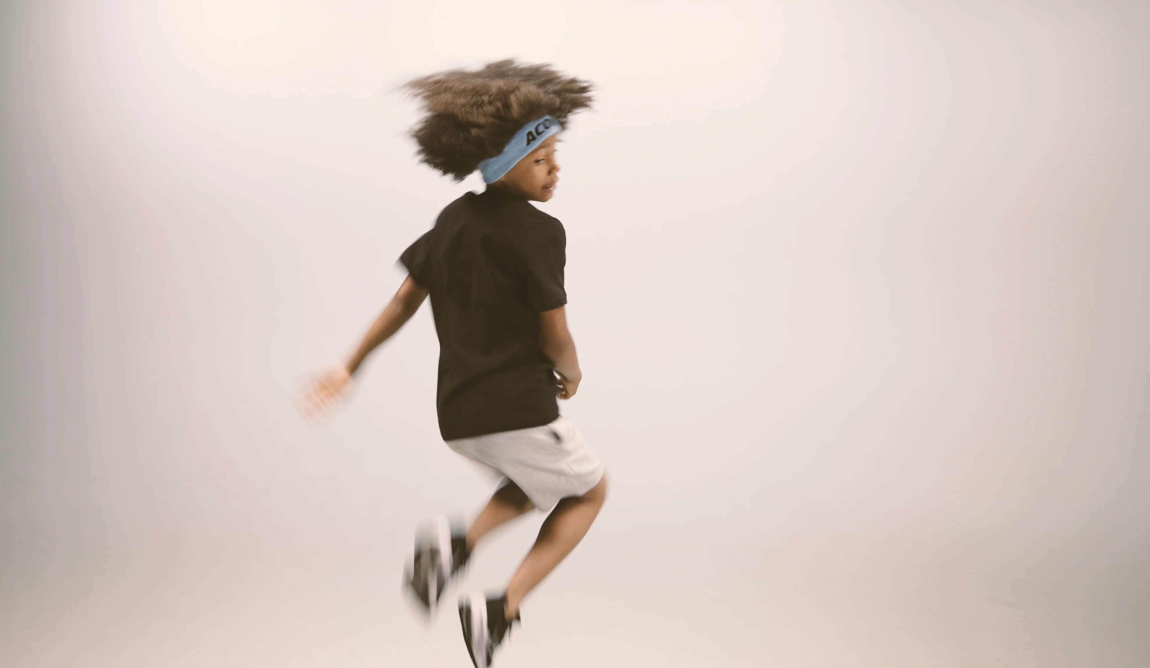 Ein springendes Kind, das ein Acon-Stirnband, ein schwarzes T-Shirt, weiße Shorts und Turnschuhe trägt. Das Bild wurde vor beigem Hintergrund aufgenommen
