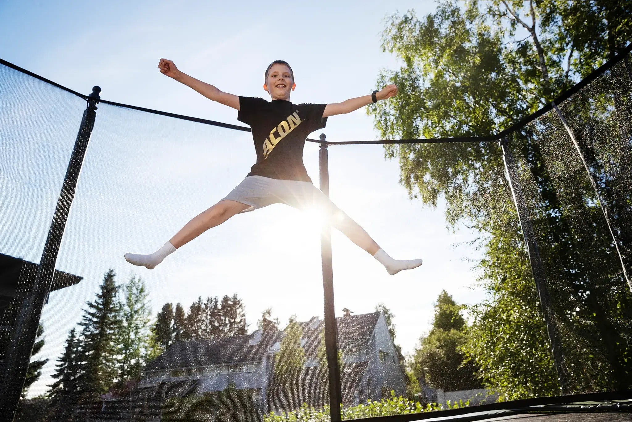 Junge mit Acon-T-Shirt beim X-Jump auf einem Acon-Trampolin, während die Sonne im Garten scheint.