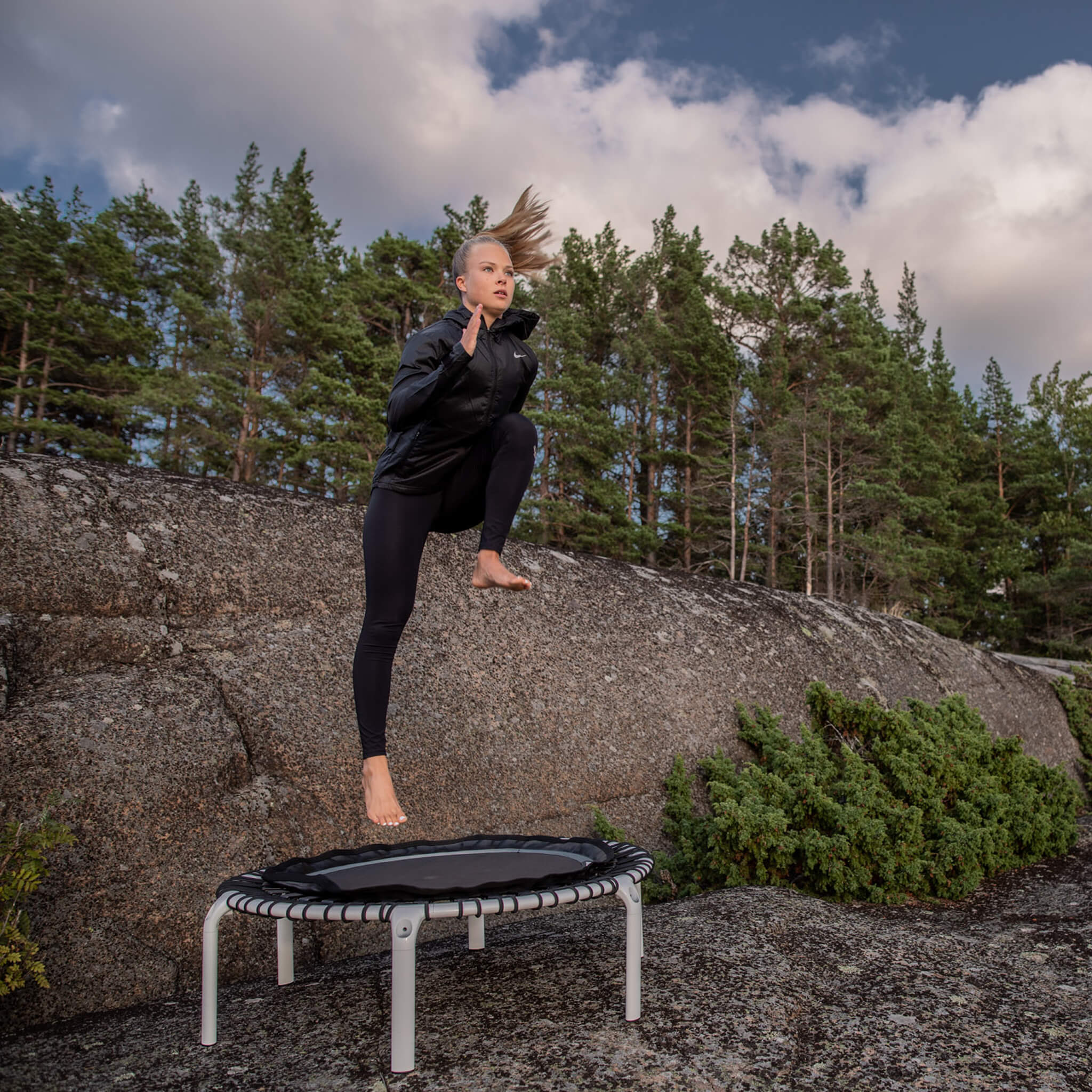 Eine Frau springt auf einem weißen Acon FIT-Trampolin in einer nordischen Waldlandschaft.