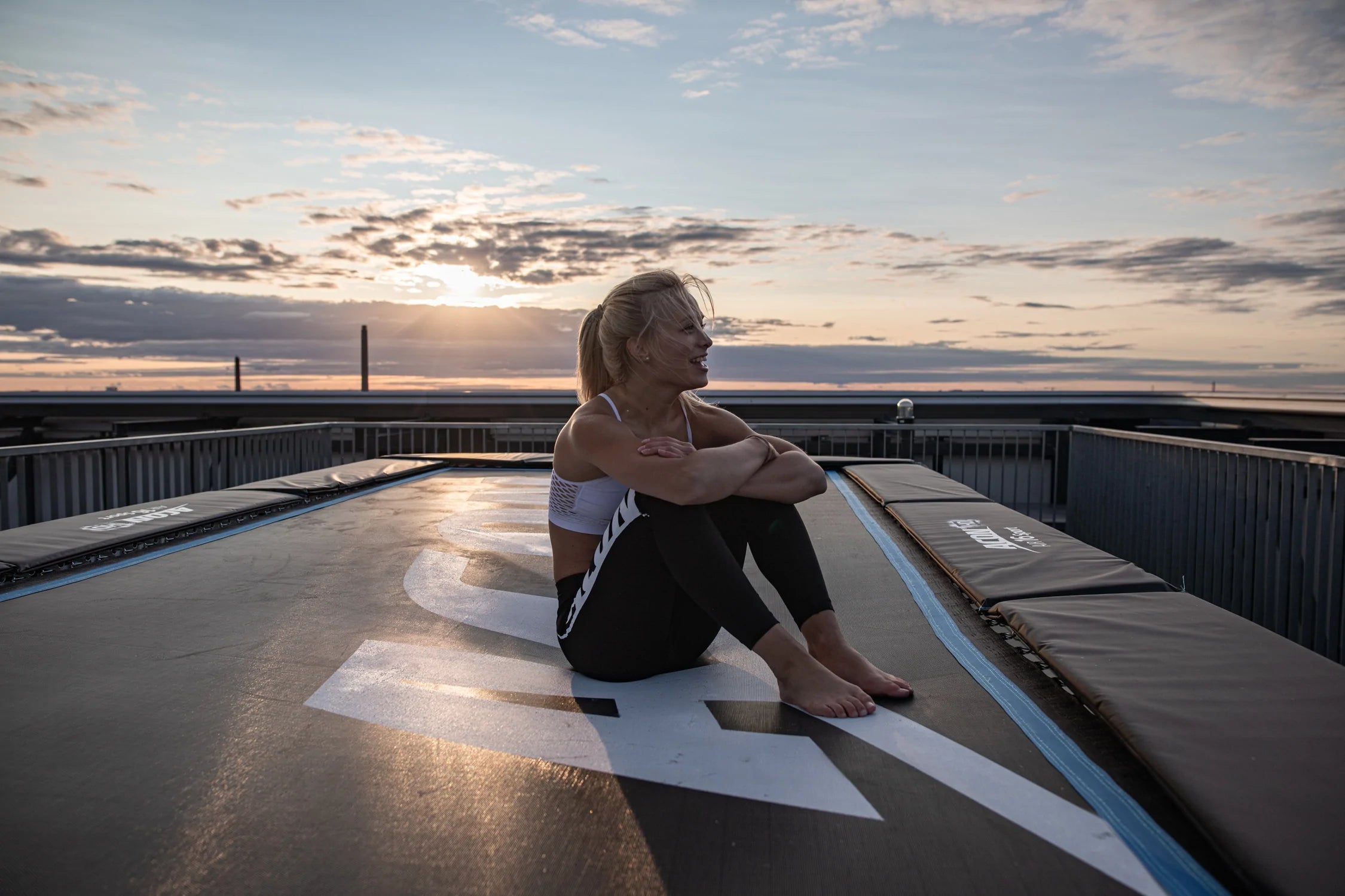 Fröhliche Frau sitzt auf einem Trampolin Acon 16 HD mit Sonnenuntergang als Hintergrund