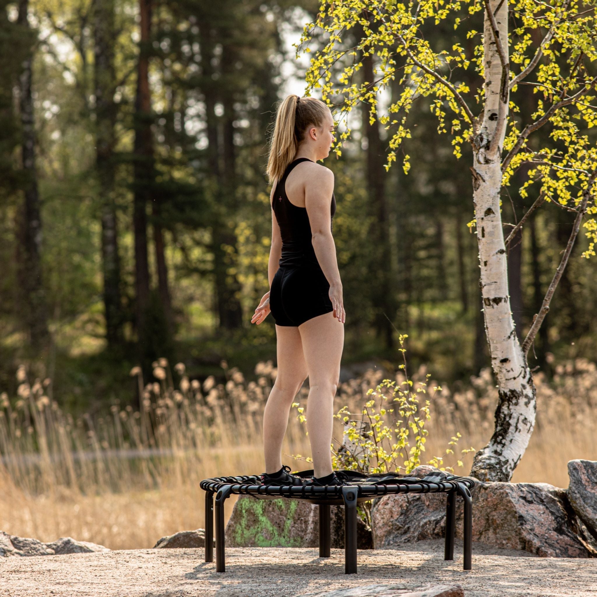 Eine Frau steht auf einer Bank im Wald, umgeben von Bäumen, mit einem Kleines Trampolin für Garten im Hintergrund.