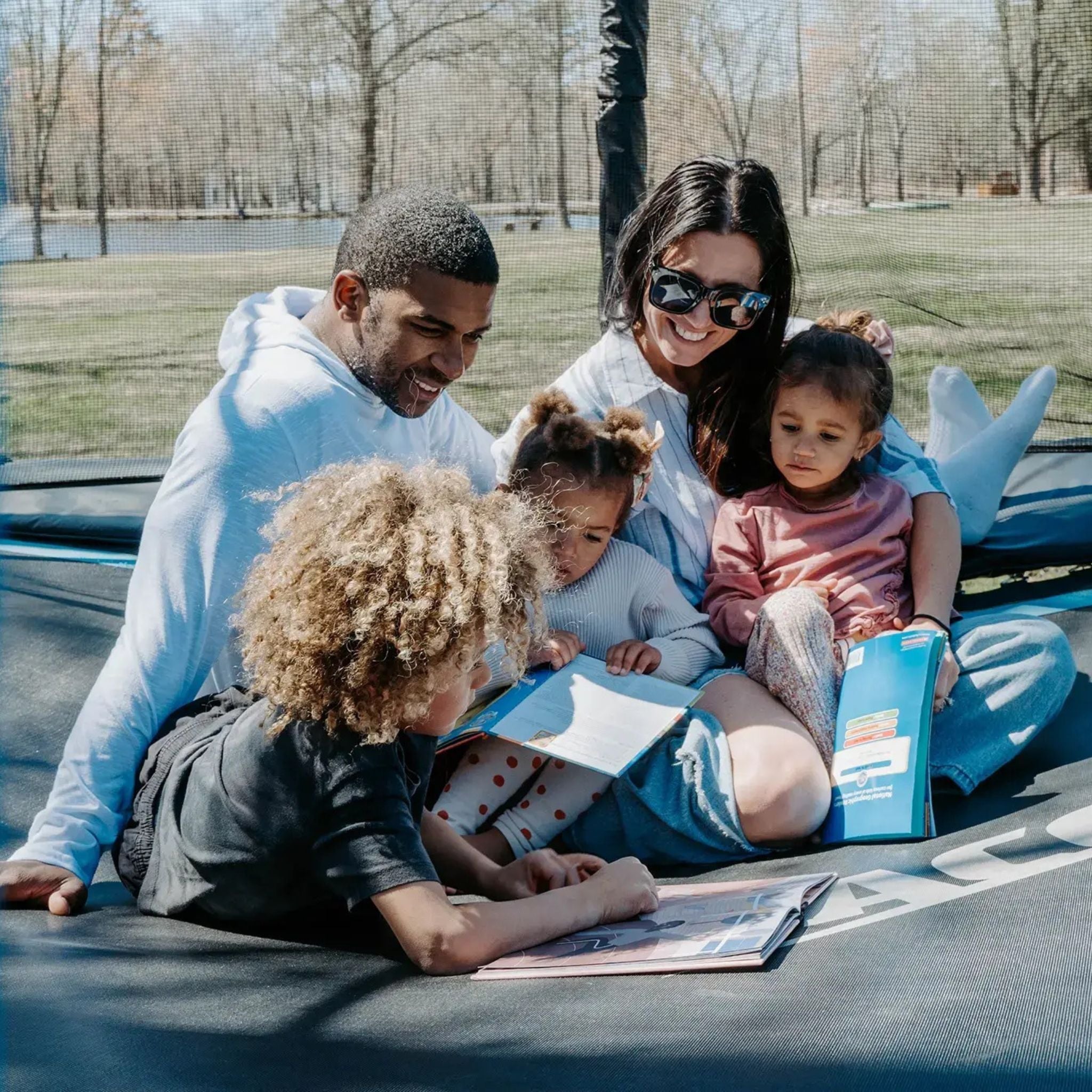 Eine Familie genießt Zeit auf einem Trampolin, während sie gemeinsam ein Buch liest, inspiriert von Trampolin Ideen. 