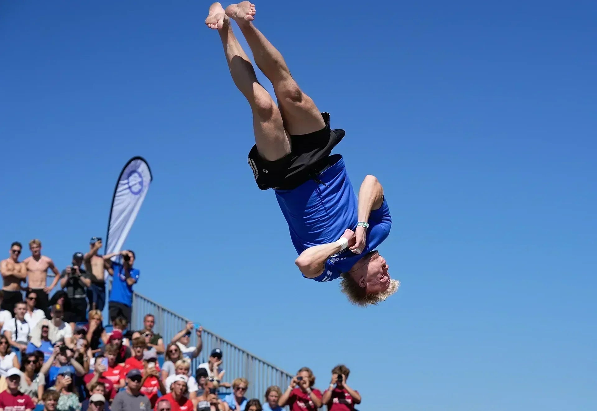 Ein männlicher Athlet vollführt einen eindrucksvollen Salto auf einem Trampolin, im Hintergrund ein blauer Himmel und eine jubelnde Menschenmenge.
