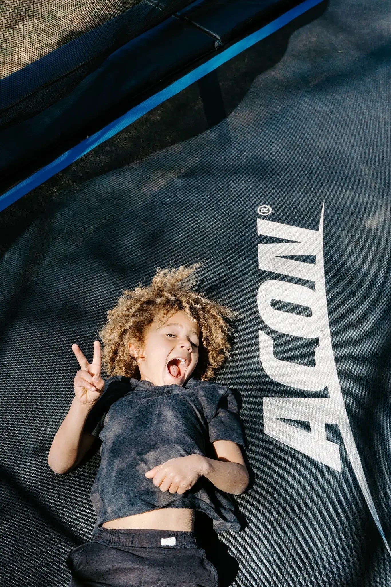 Junges Kind liegt auf einem Acon-Trampolin, zeigt das Peace-Zeichen und lächelt.