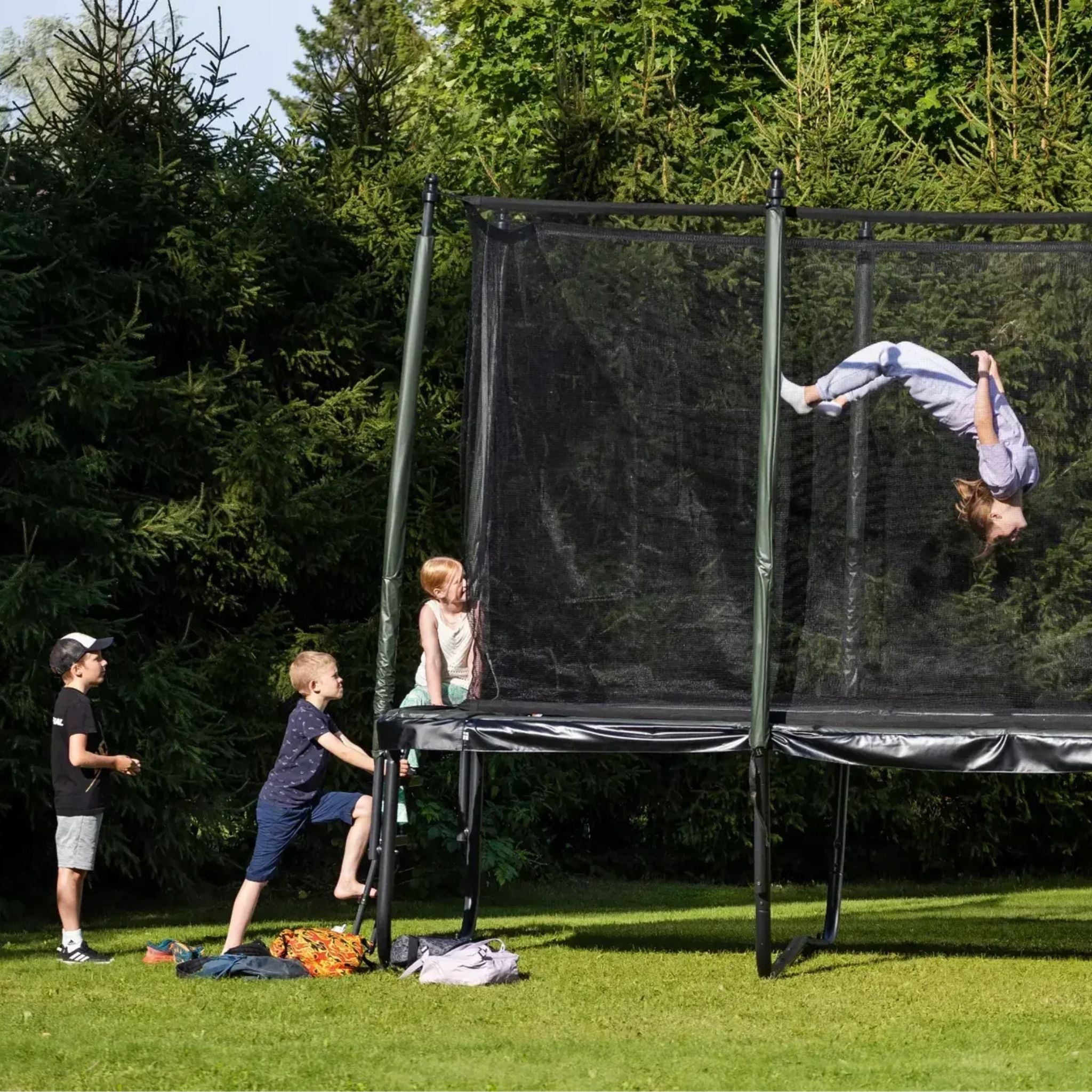 Gruppe von Kindern, die auf einem Trampolin mit Sicherheitsnetz im Freien spielen – ein Kind macht einen Überschlag in der Luft, während die anderen auf ihre Reihe warten.