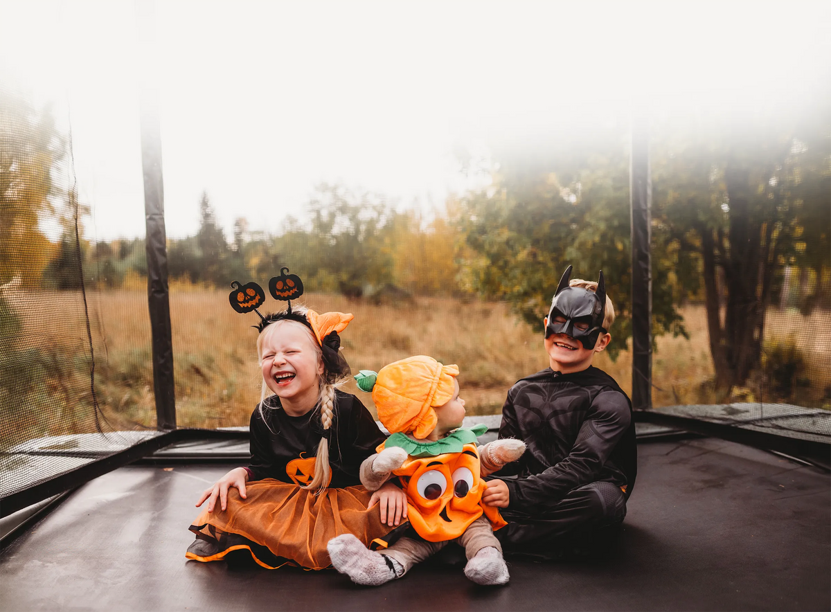Drei Kinder in Halloween-Kostümen sitzen gemeinsam auf einem ACON 16HD Trampolin.