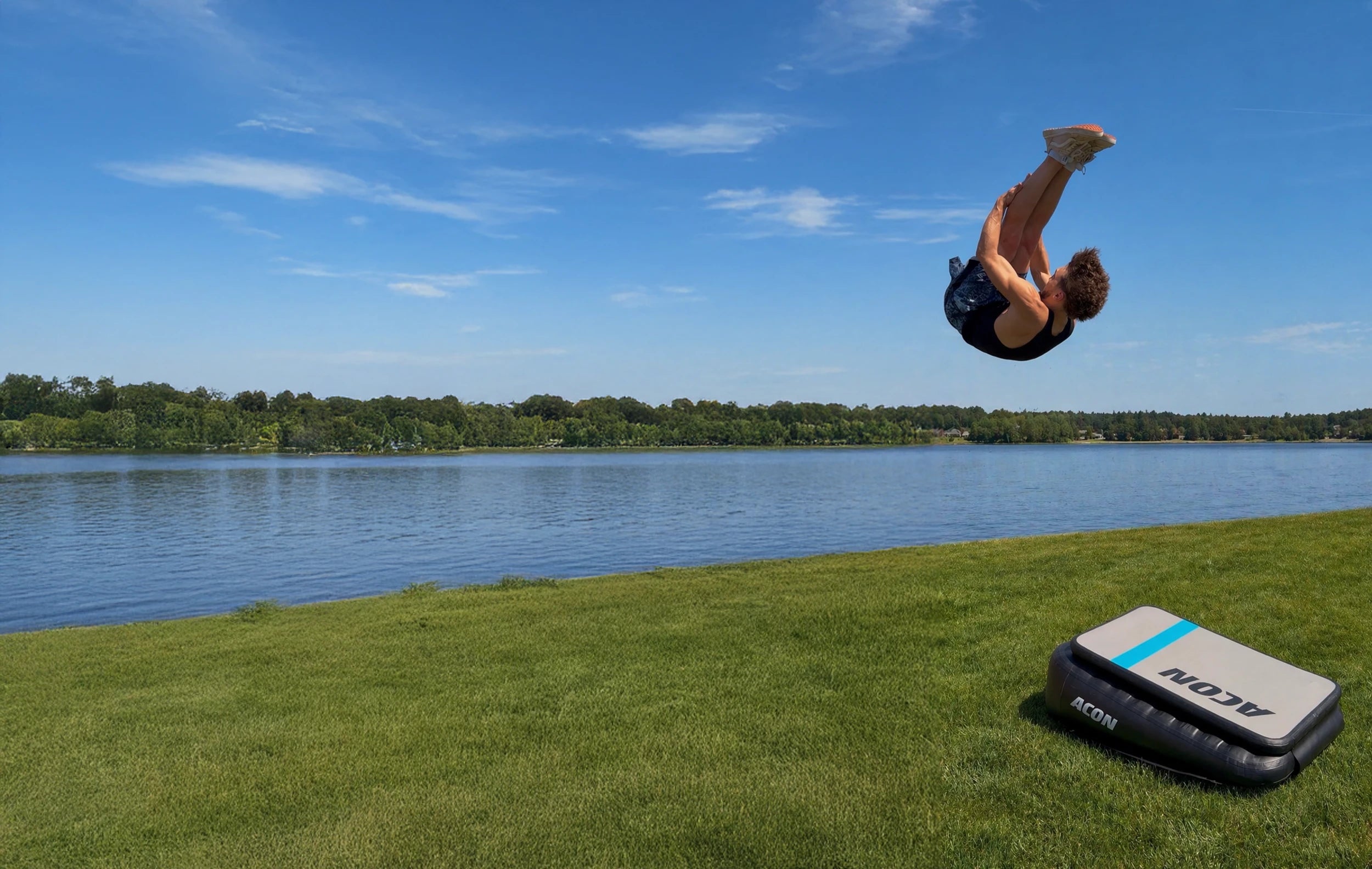 Ein Mann springt einen Pike Front auf einem Acon Airboard am Flussufer, das Airboard steht auf Gras.