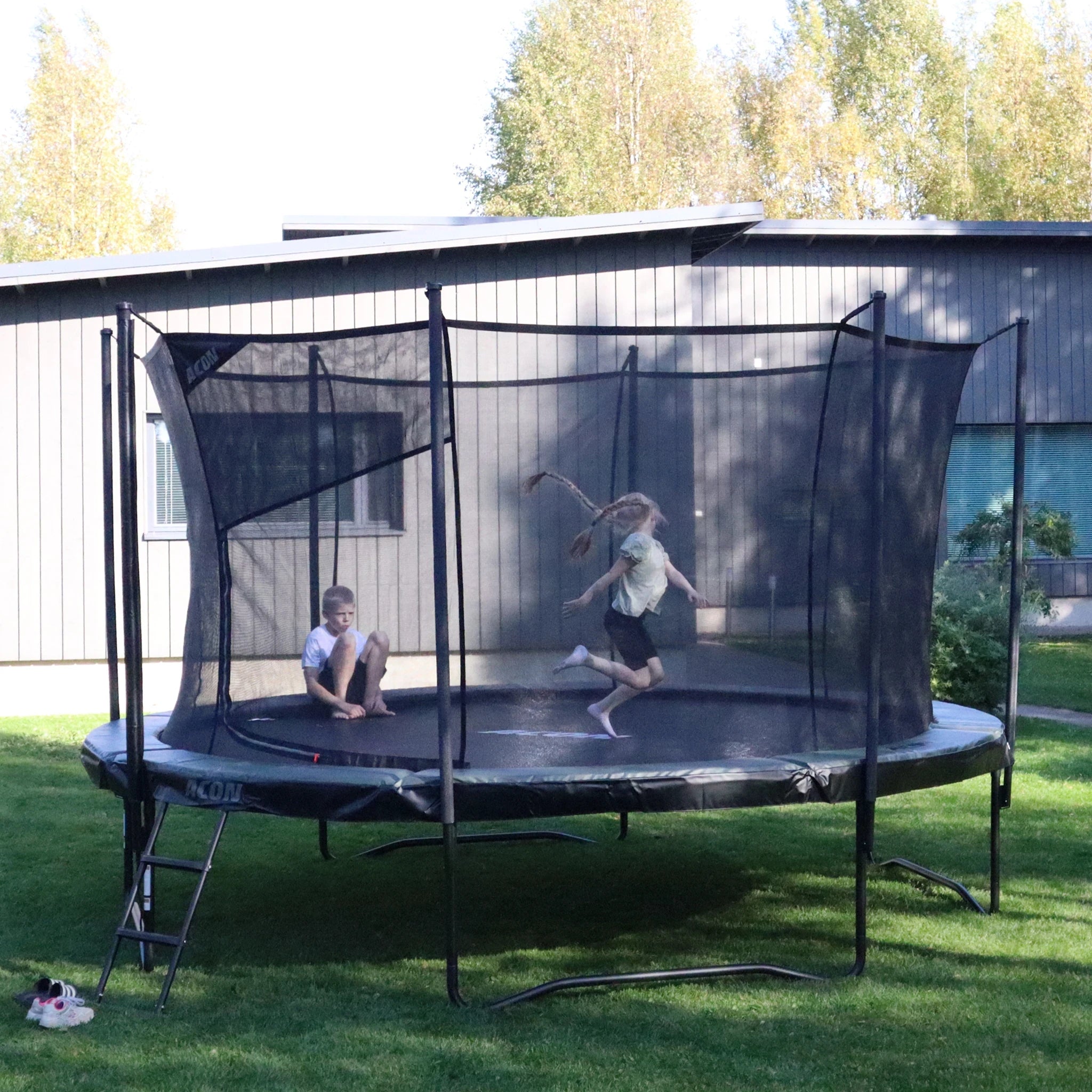 Acon Air-Trampolin im Garten eines Einfamilienhauses. Ein Junge sitzt auf dem Trampolin, ein Mädchen springt.