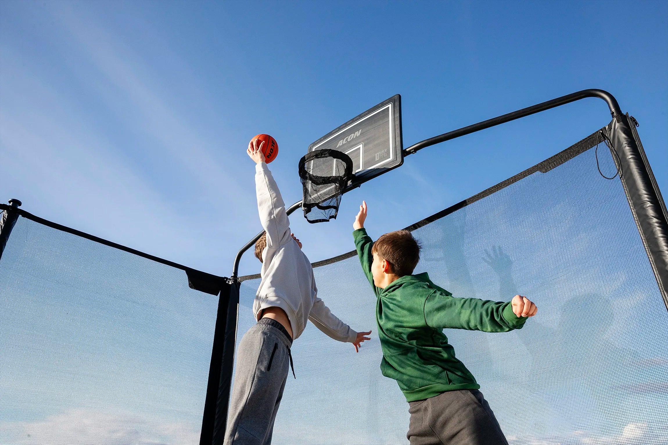 Zwei Personen spielen Basketball auf einem Acon X Trampolin im Freien unter klarem blauen Himmel.