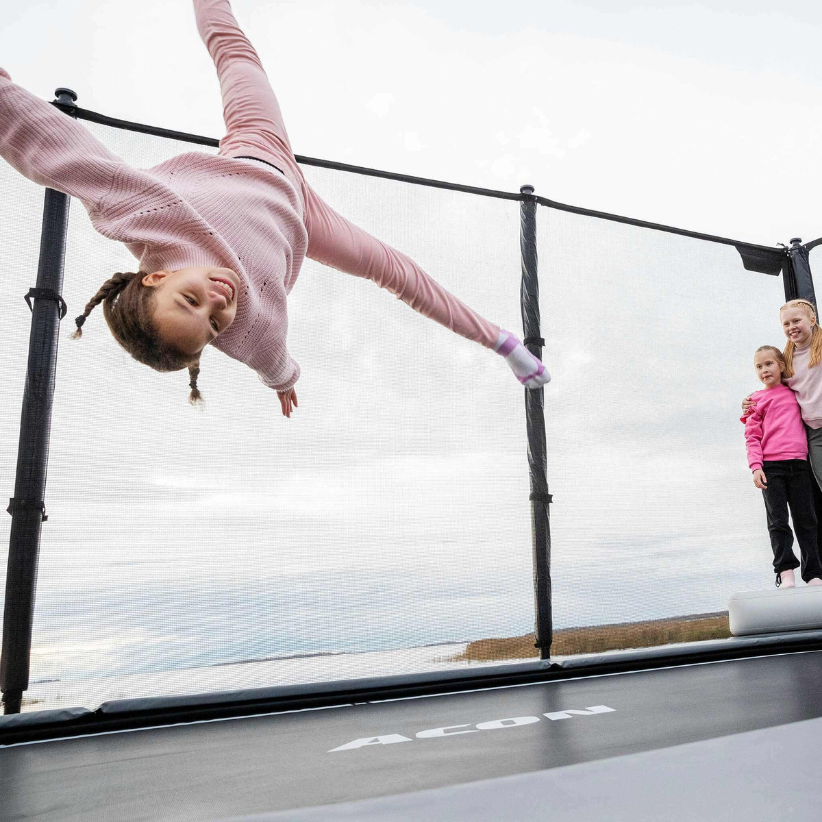 Ein Mädchen springt auf dem Acon X Trampolin, zwei Mädchen stehen daneben und schauen zu.