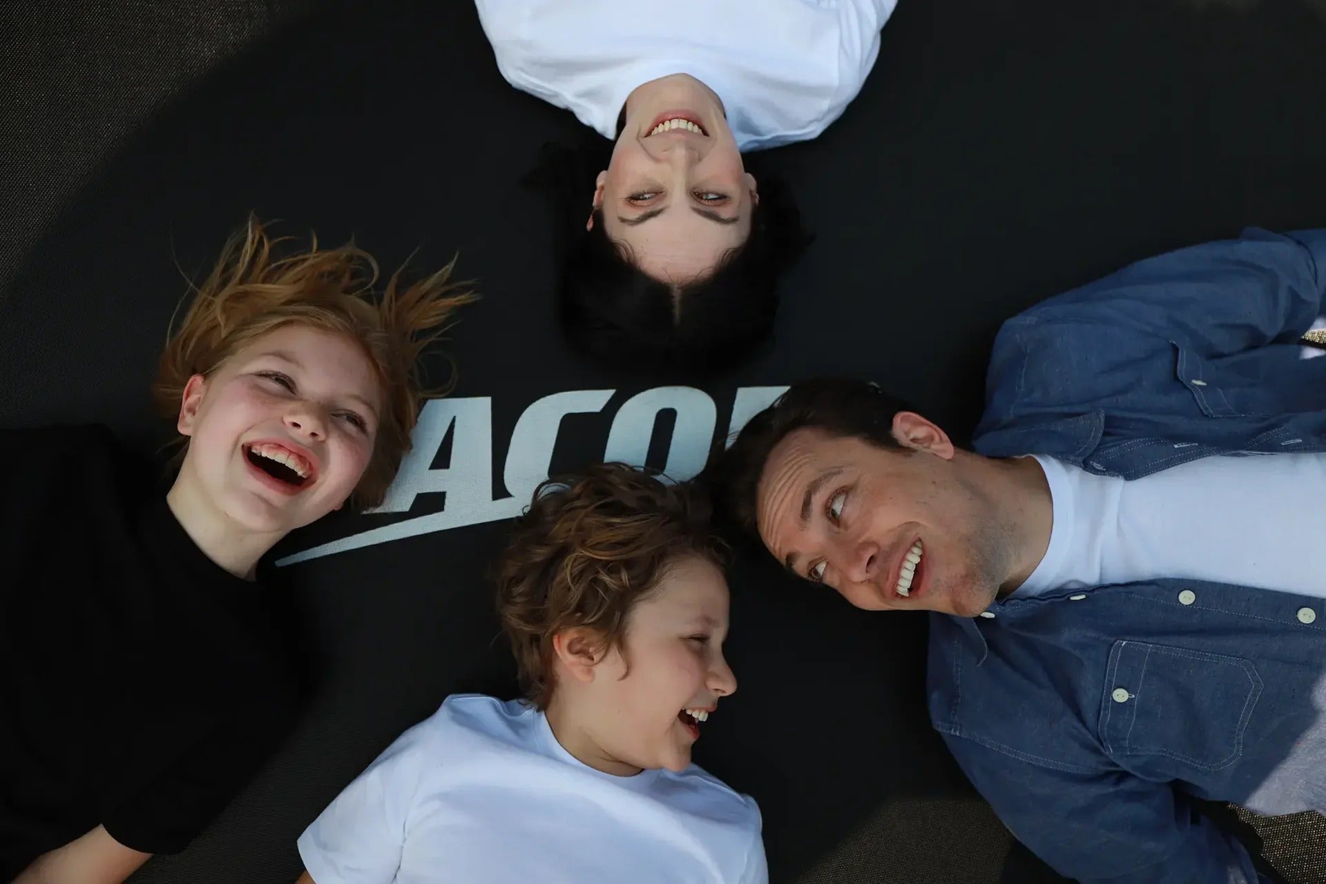Nahaufnahmefoto einer vierköpfigen Familie, die lächelnd auf einem Acon-Trampolin liegt.