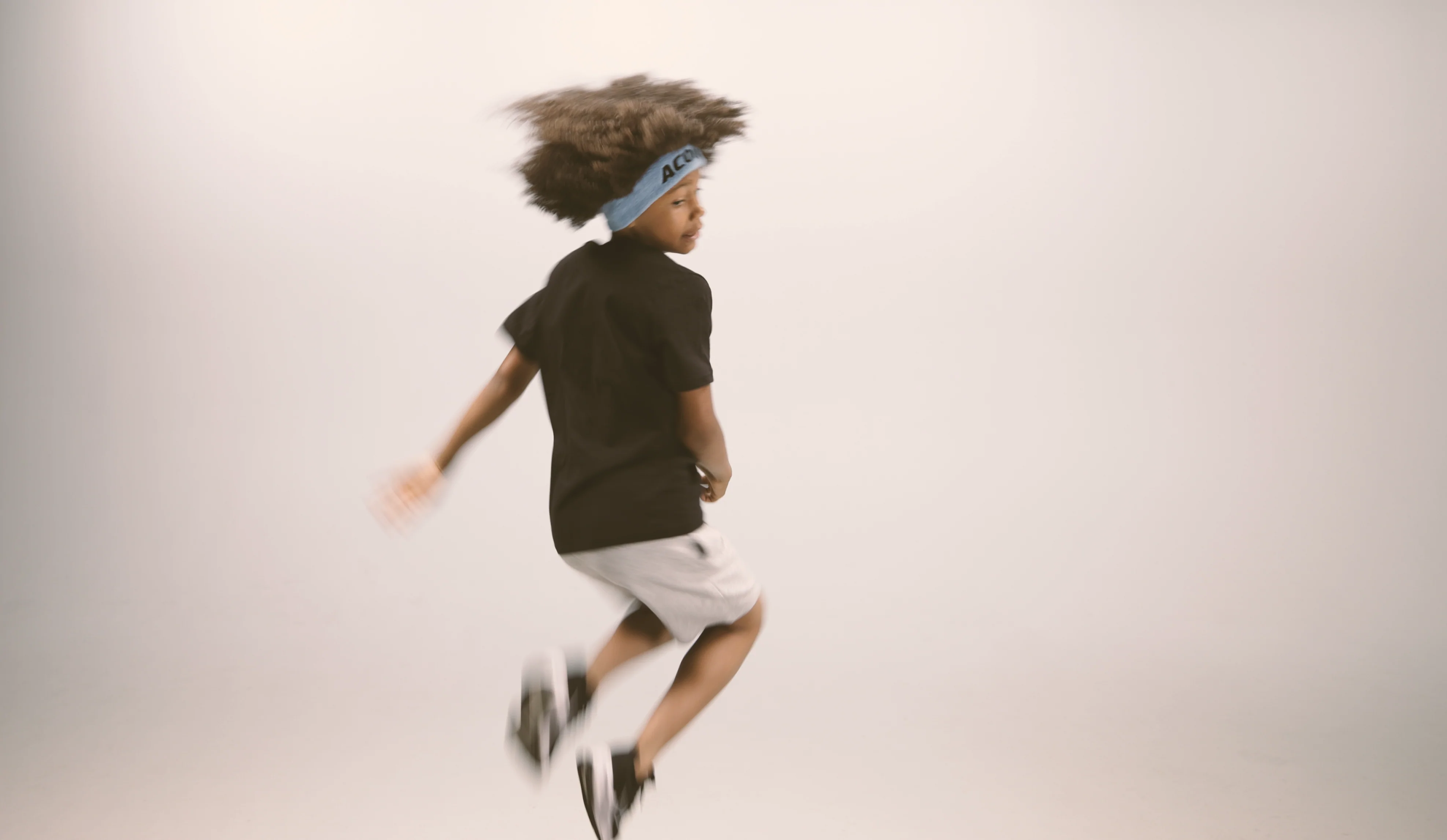 Ein springendes Kind, das ein Acon-Stirnband, ein schwarzes T-Shirt, weiße Shorts und Turnschuhe trägt. Das Bild wurde vor beigem Hintergrund aufgenommen 