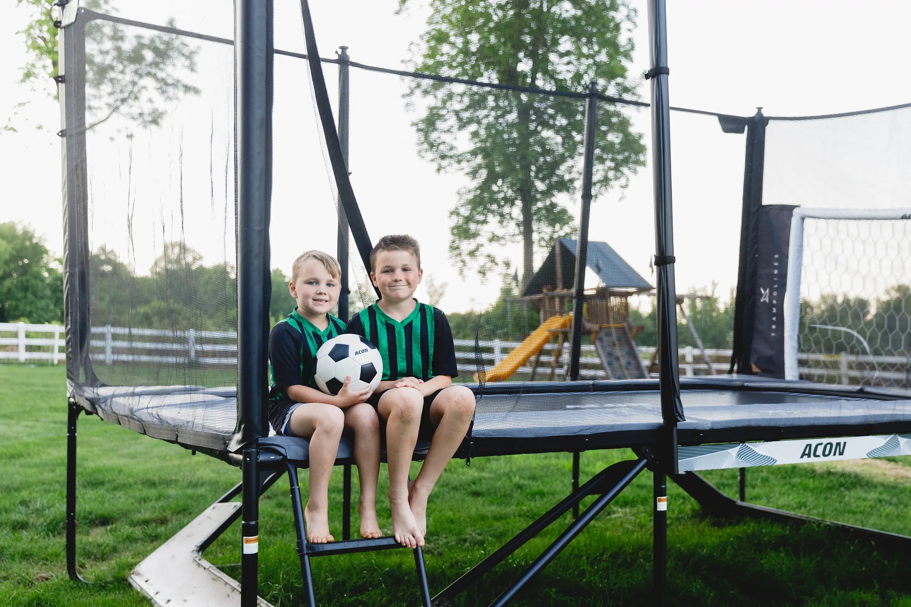 Zwei Kinder mit Fußball auf Acon Trampolin im Garten mit Sicherheitsnetz und Trampolin-Fußballtor.