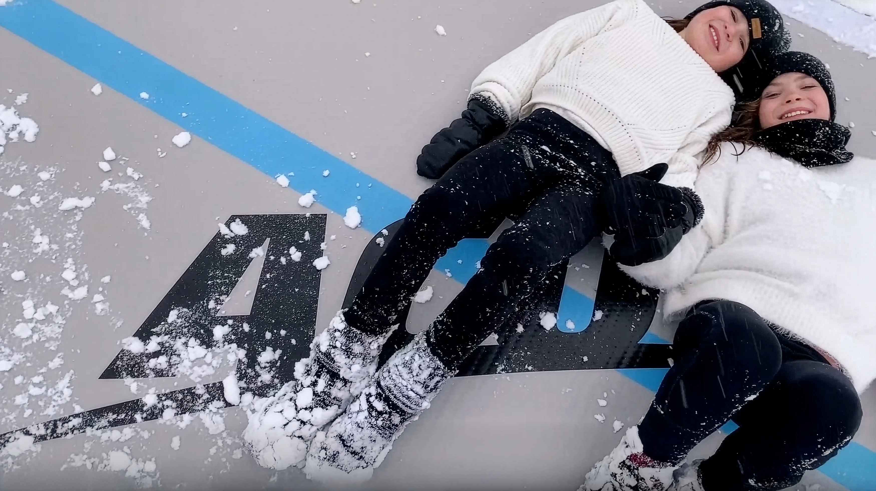 Zwei lächelnde Schwestern liegen nebeneinander auf einer Acon-Airtrack-Matte, auf der etwas Schnee liegt.