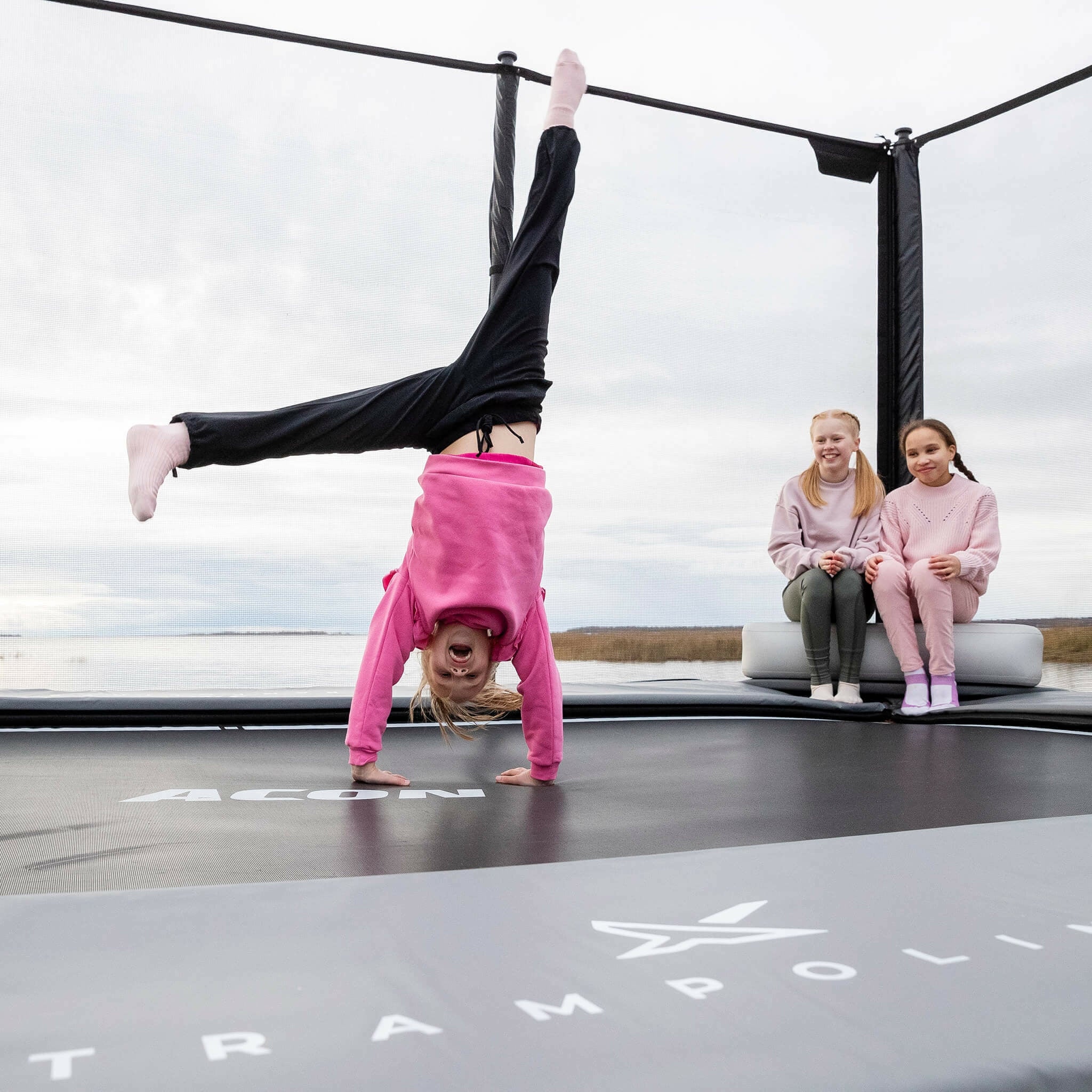 Zwei Mädchen sitzen auf dem aufblasbaren Acon X Triangle Block. Das Mädchen steht mit den Händen auf dem Trampolin.