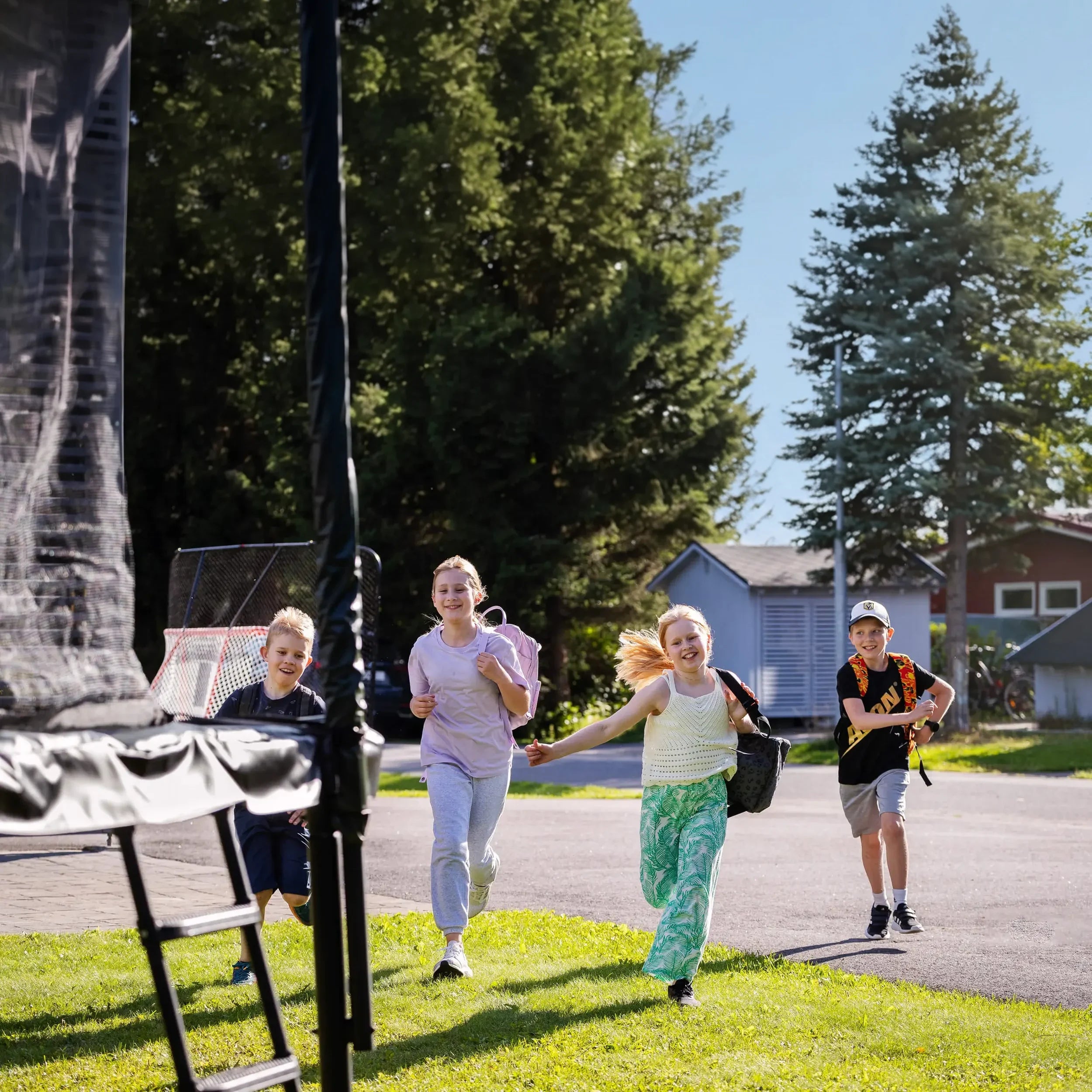 Vier Kinder laufen nach der Schule mit Rucksäcken zu einem Acon Trampolin im sonnigen Garten.