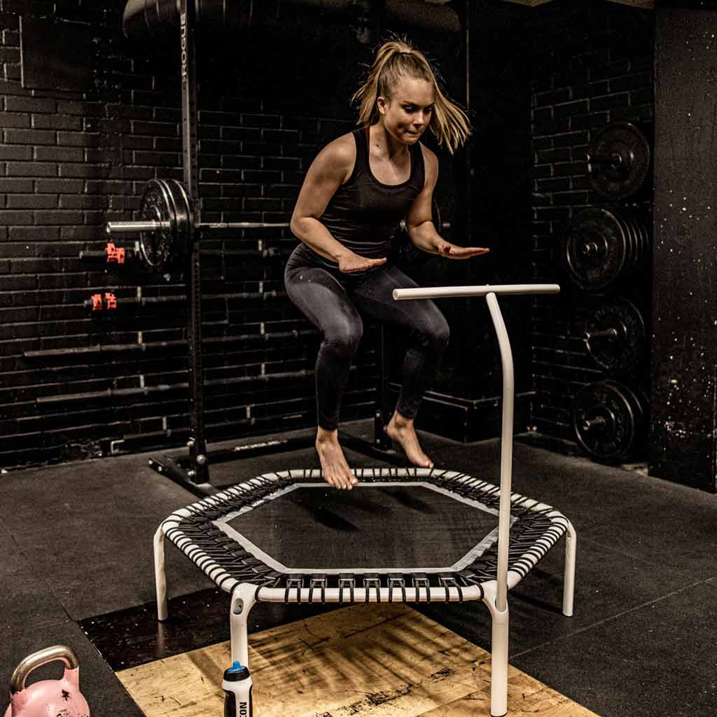 Eine Frau, die mit einem weißen ACON FIT-Sechskant-Trampolin mit Griffstange in einer Turnhalle trainiert