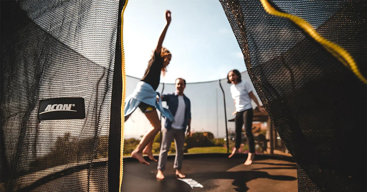 Familie genießt Spaß im Freien auf einem Acon-Trampolin, gesehen durch den geöffneten Eingang des Sicherheitsnetzes.