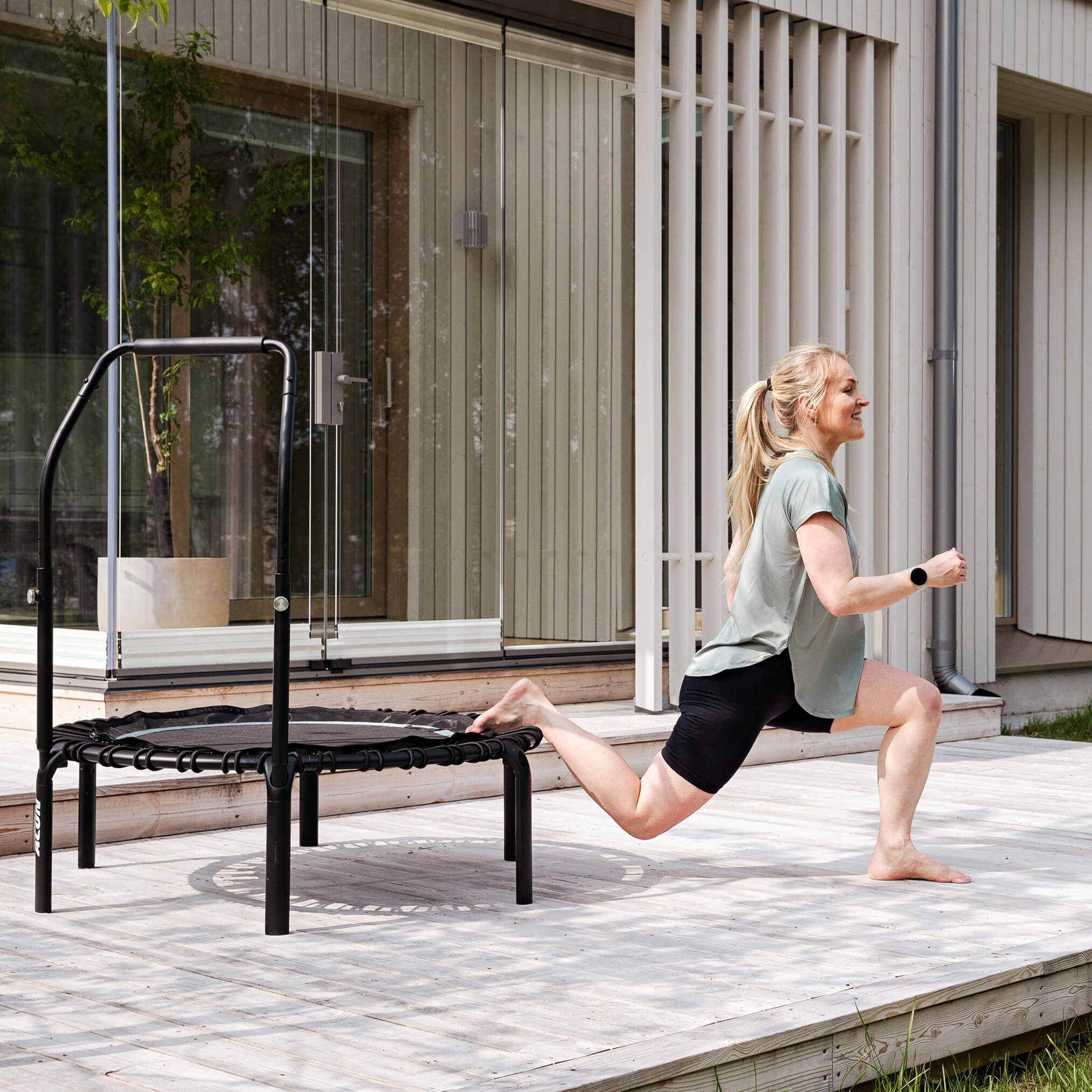 Eine Frau macht einbeinige Kniebeugen auf einem schwarzen Acon FIT-Trampolin auf einer Terrasse.