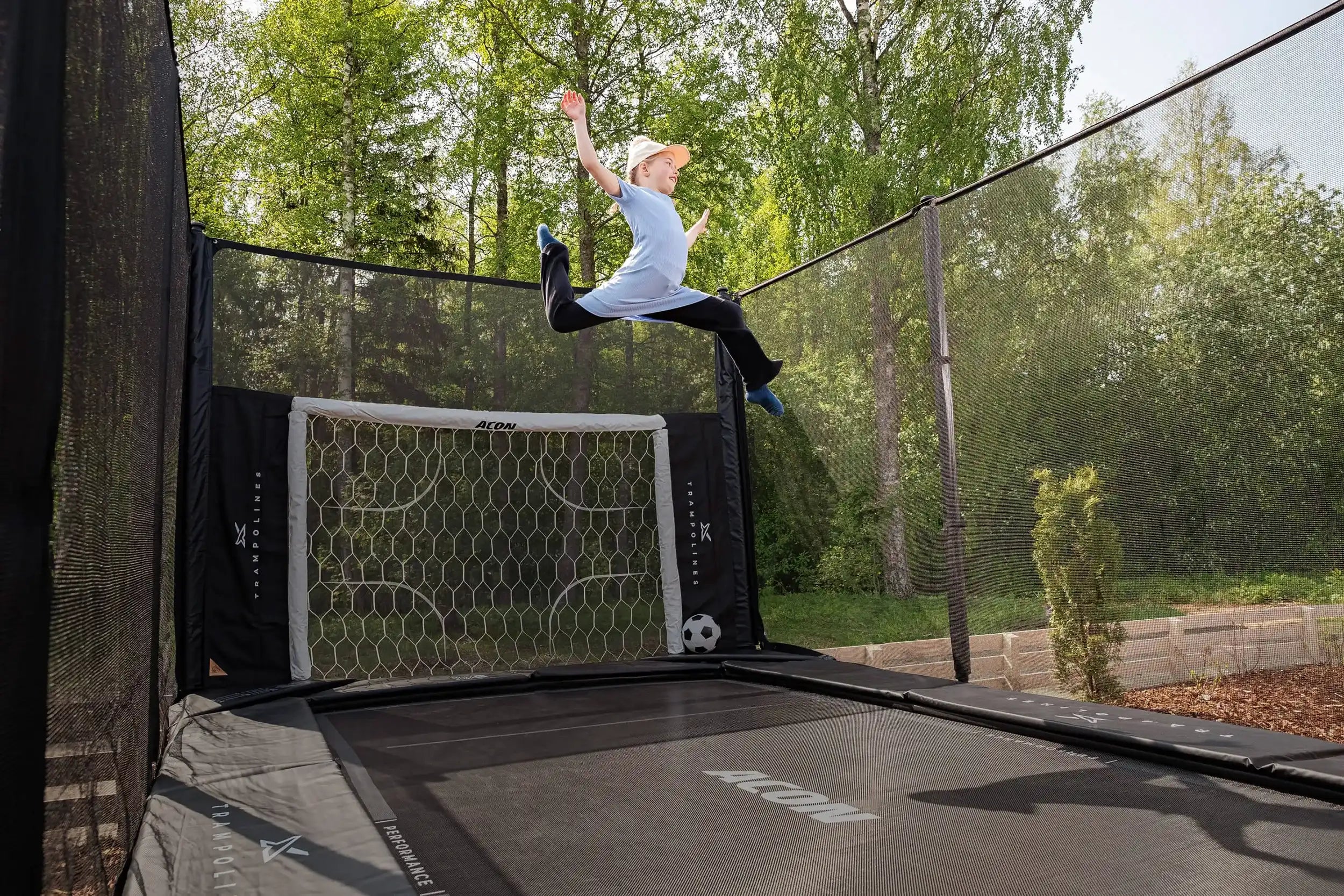 Mädchen springt auf einem Acon X Trampolin im sommerlichen Garten, ausgestattet mit einem Acon Fußballtor.