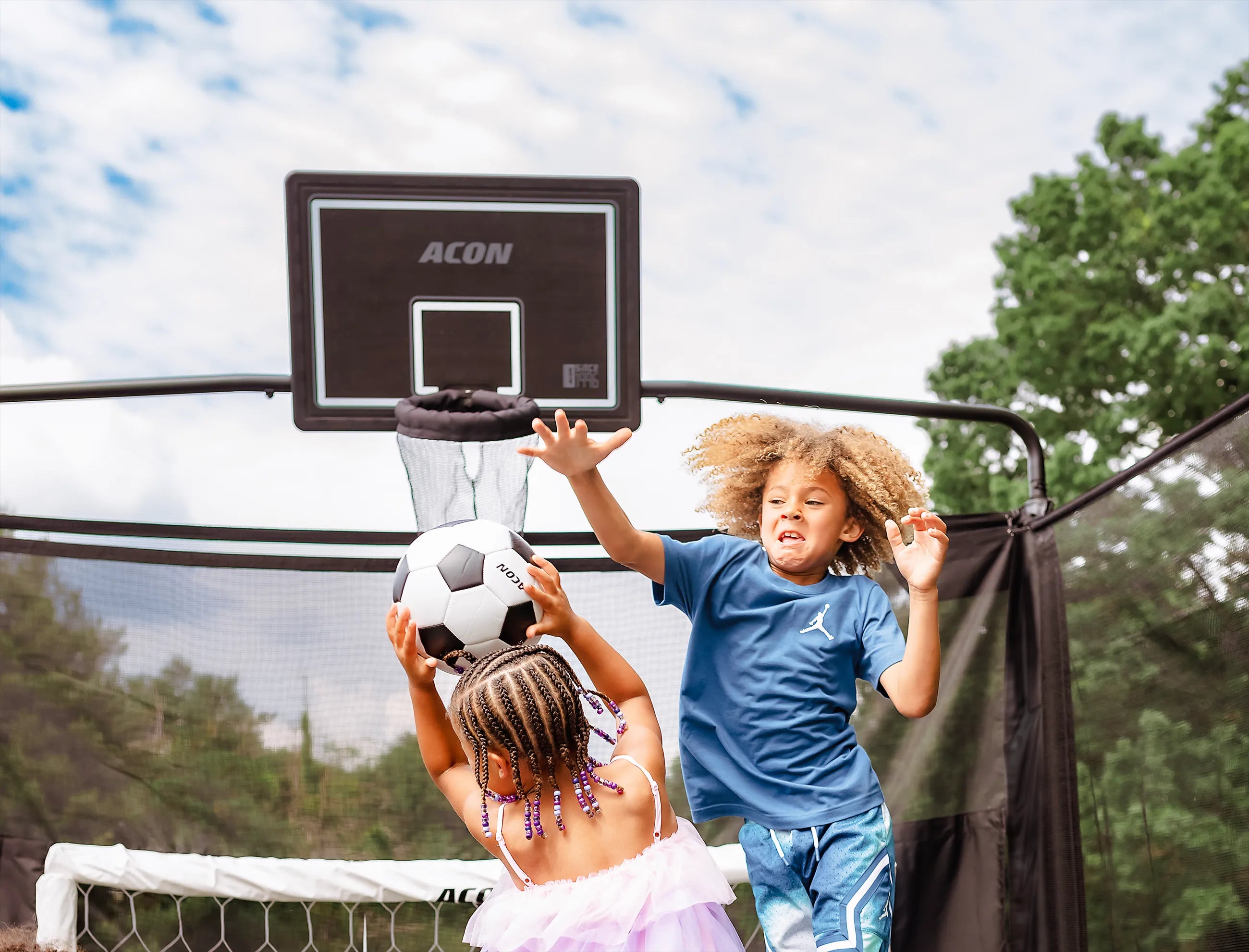 Zwei Kinder spielen mit einem Fußball auf einem Acon X Trampolin mit Fußballtor und Basketballkorb.