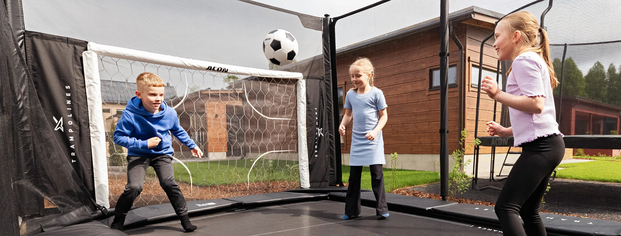 Drei Kinder spielen Fußball auf einem Acon X-Trampolin im Garten eines Einfamilienhauses. Am Ende des Trampolins befindet sich ein Fußballtor-Zubehör für das Acon X-Trampolin, auf das die Kinder ihre Schüsse richten.